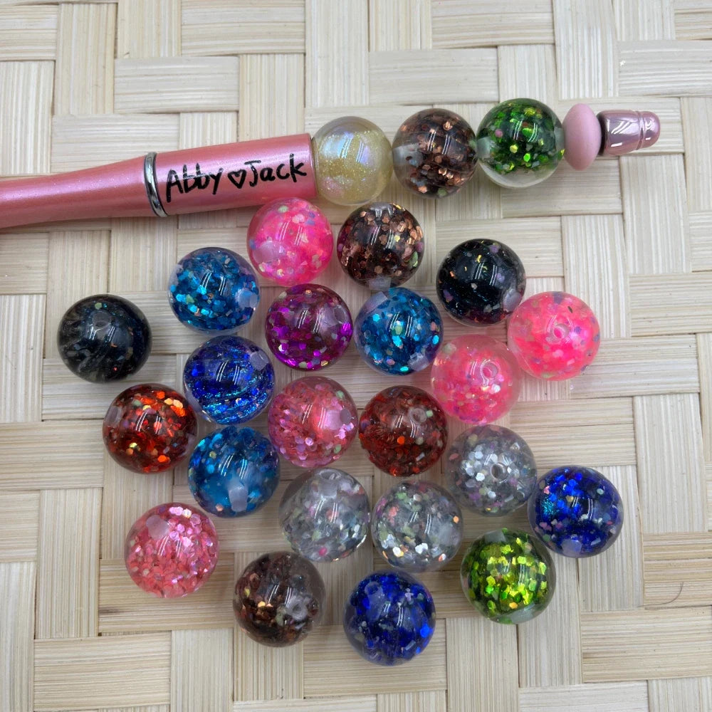 16MM glitter confetti acrylic beads in assorted colors including pink, blue, black, green, and red with chunky glitter fill, displayed next to a pink beadable pen on a woven surface.