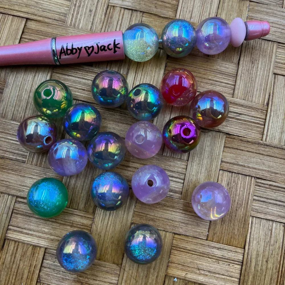 16MM aurora AB iridescent acrylic beads in rainbow holographic purple green pink and multicolor finishes displayed on a woven surface next to a beadable pen