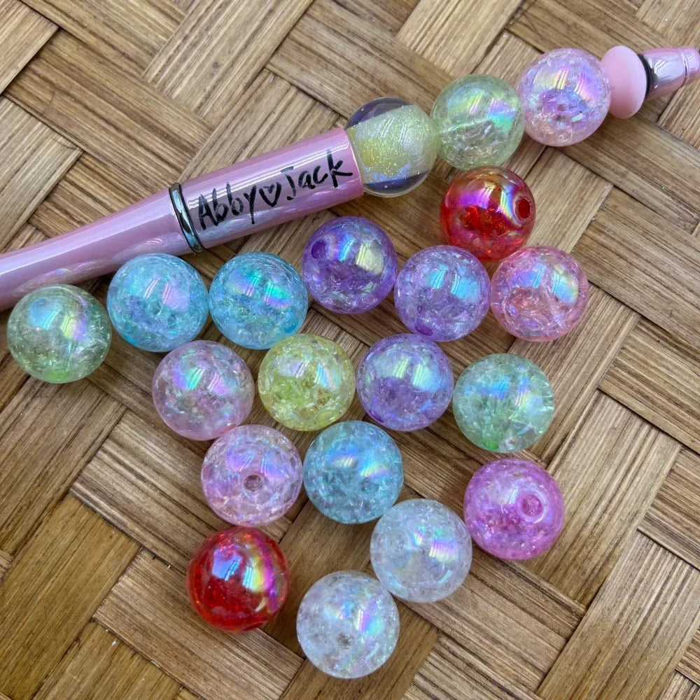 16MM aurora iridescent crackle acrylic beads in assorted pastel and bold colors including pink, purple, teal, yellow, red, and clear, displayed next to a pink beadable pen on a woven surface.