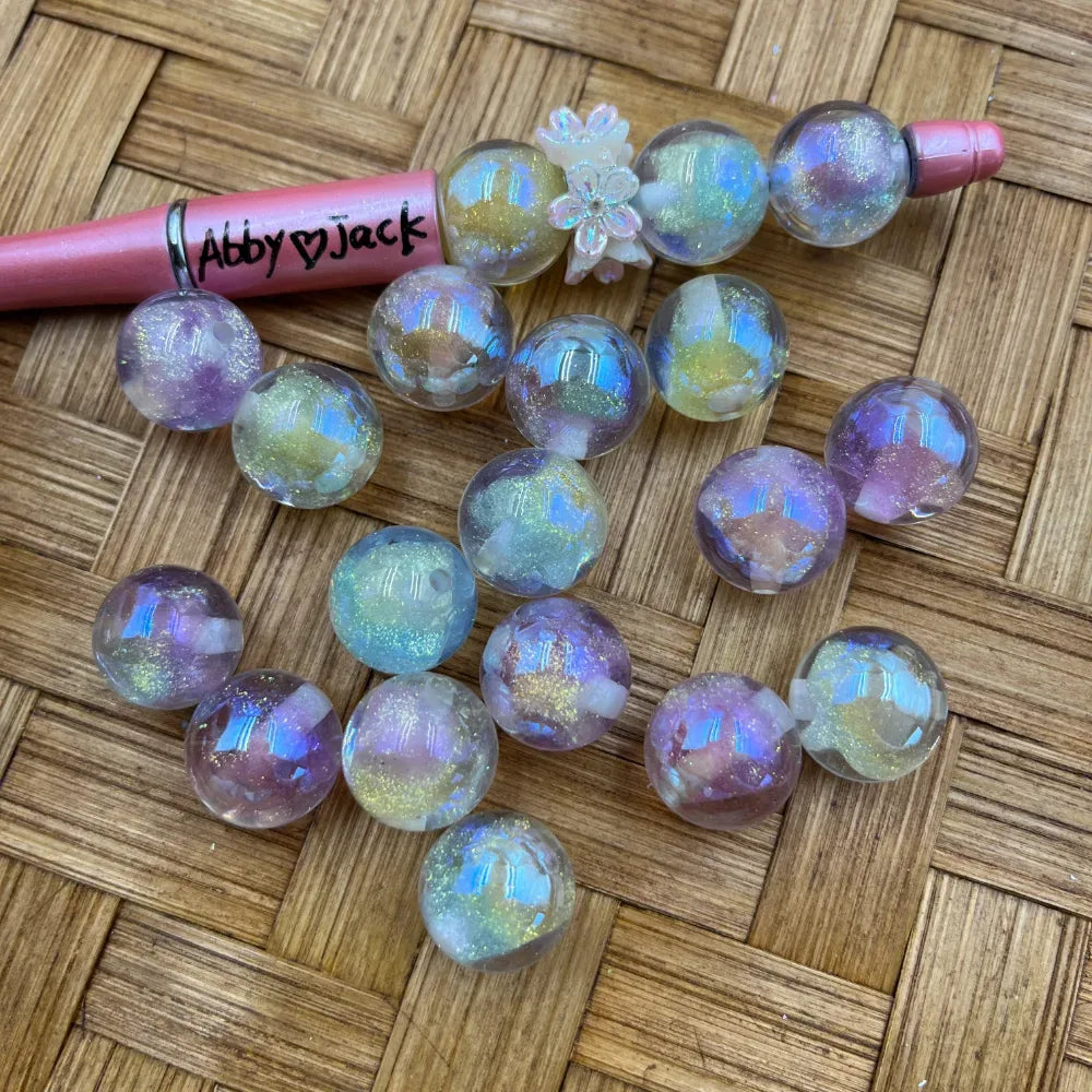 16MM aurora galaxy glitter acrylic beads with iridescent holographic shimmer in purple, green, and blue tones, displayed next to a beadable pen on a wicker surface