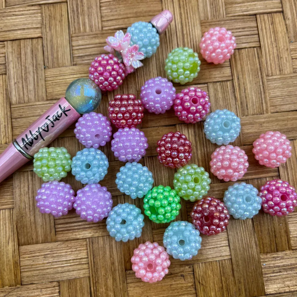 16MM berry bumpy textured acrylic beads in pastel AB iridescent colors including pink, purple, light blue, green, and hot pink arranged on a woven surface next to a beadable pen