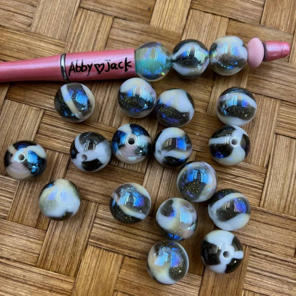16MM black and white cow print acrylic beads with iridescent blue galaxy glitter finish, shown next to a pink beadable pen on a wicker surface