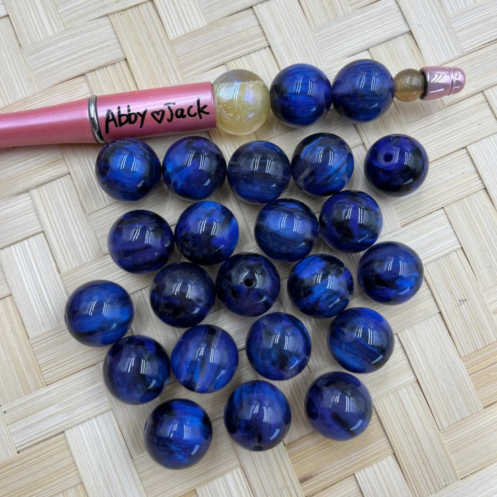 16MM deep blue galaxy swirl acrylic beads with black marbled pattern displayed on wicker surface next to a beadable pen for size reference.