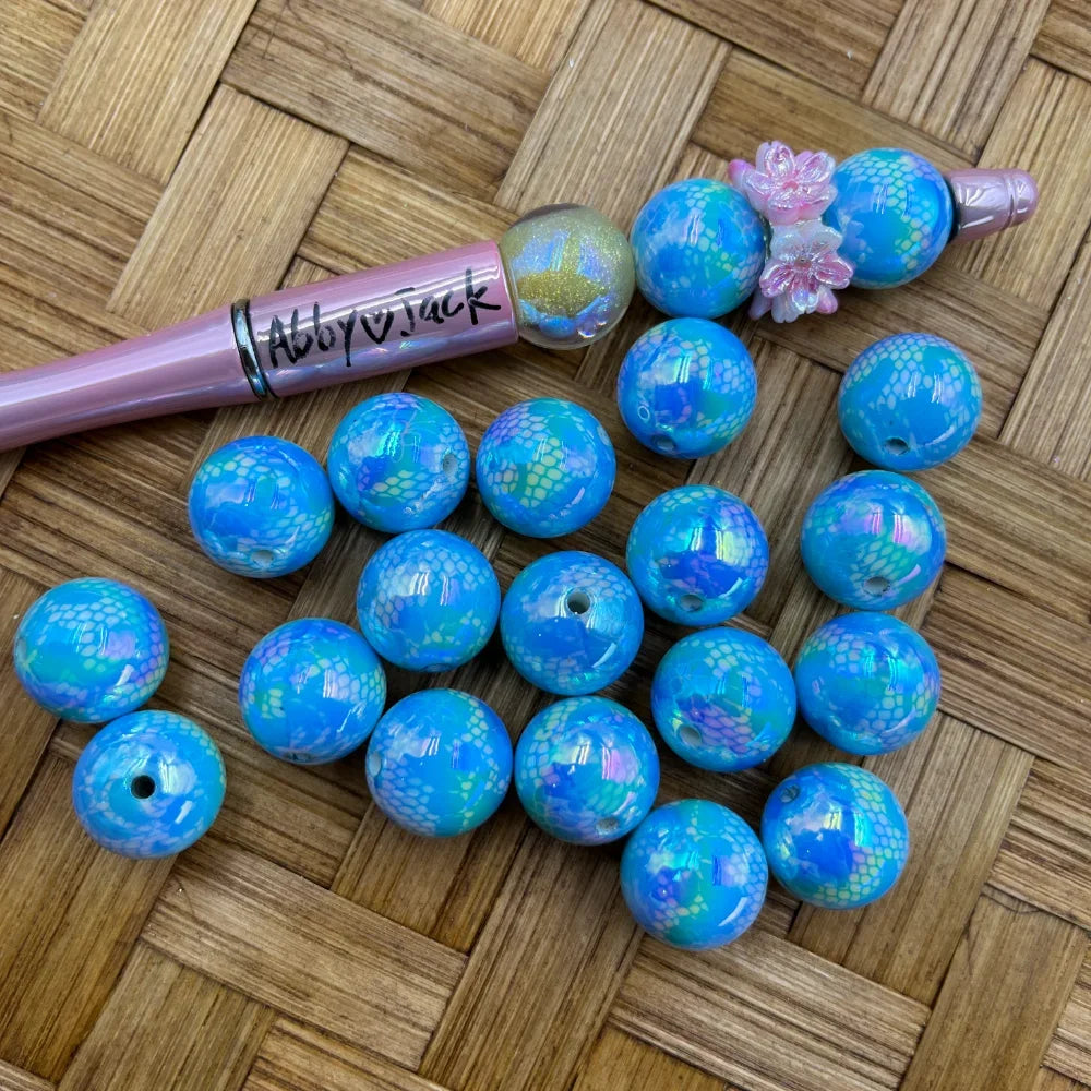 16MM blue iridescent mermaid scale patterned acrylic beads displayed on a woven surface next to a pink beadable pen decorated with matching beads and pink flower embellishments.