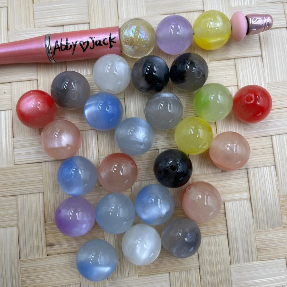 16MM cat eye acrylic beads in assorted colors including red, blue, black, yellow, pink, lavender, and gray displayed next to a beadable pen on a woven surface.
