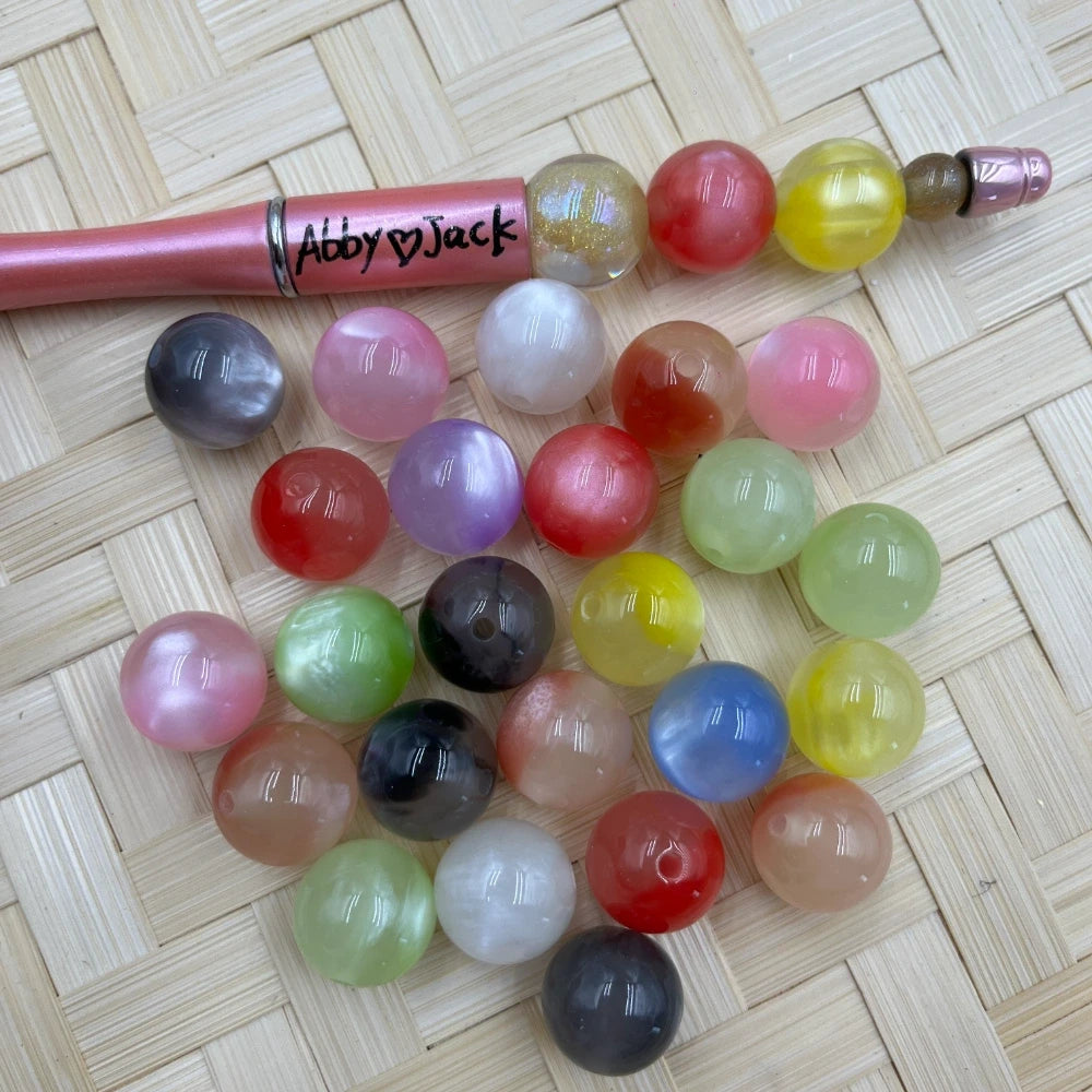 16MM mixed color cat eye acrylic beads in assorted colors including red, pink, green, blue, yellow, white, gray, and purple, displayed next to a beadable pen on a woven surface.