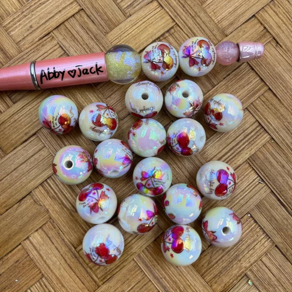 16MM white iridescent acrylic beads with cherry and butterfly print pattern, holographic shimmer finish, shown with a pink beadable pen on a wicker surface
