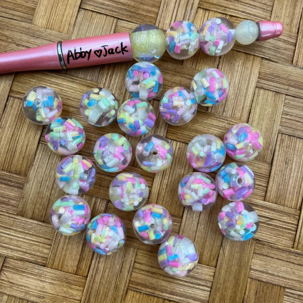 16MM clear acrylic round beads filled with pastel rainbow sprinkle confetti pieces in pink, blue, yellow, purple and white, displayed on a wicker surface next to a pink beadable pen.