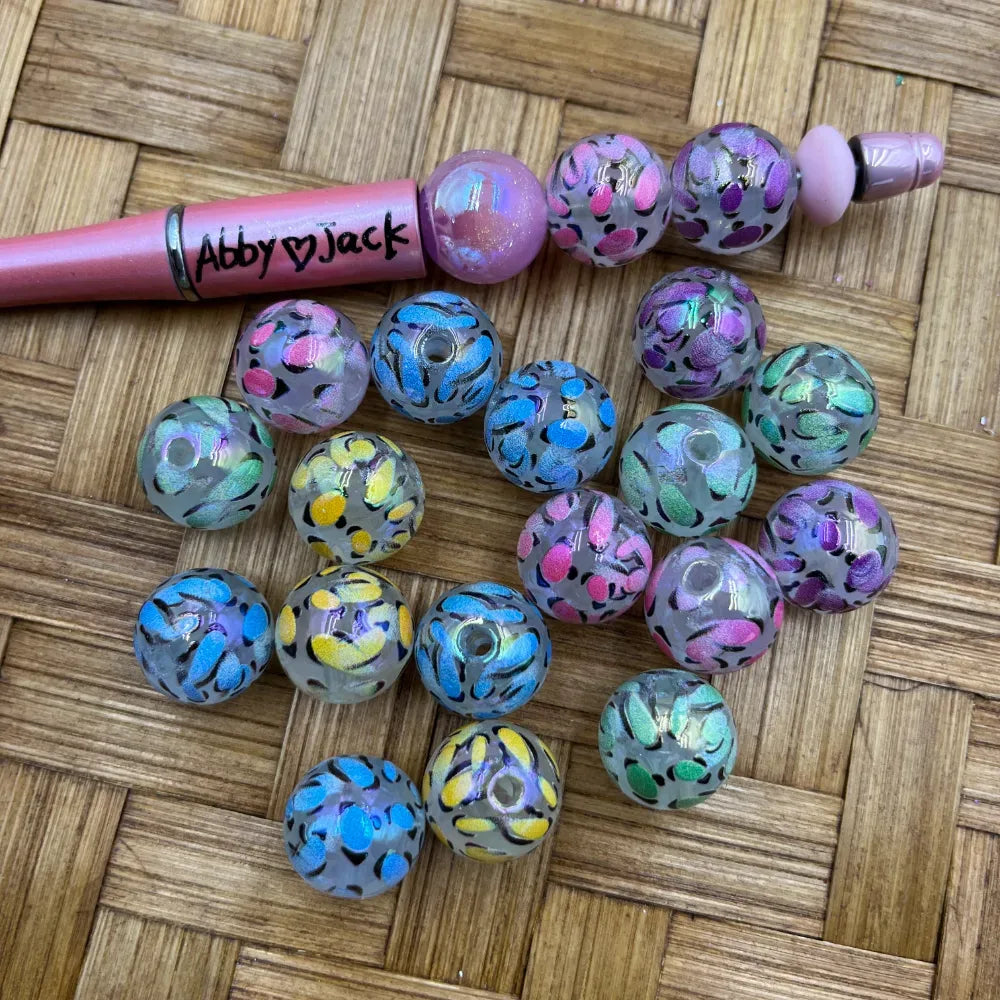 16MM clear acrylic beads with iridescent multicolor leopard print pattern in pink, blue, yellow, purple and green, displayed next to a pink beadable pen on a woven surface.