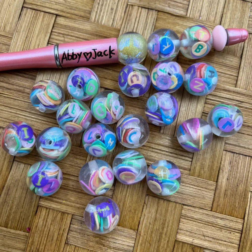 16MM clear acrylic round beads filled with colorful pastel alphabet letter confetti pieces, shown next to a pink beadable pen on a woven surface