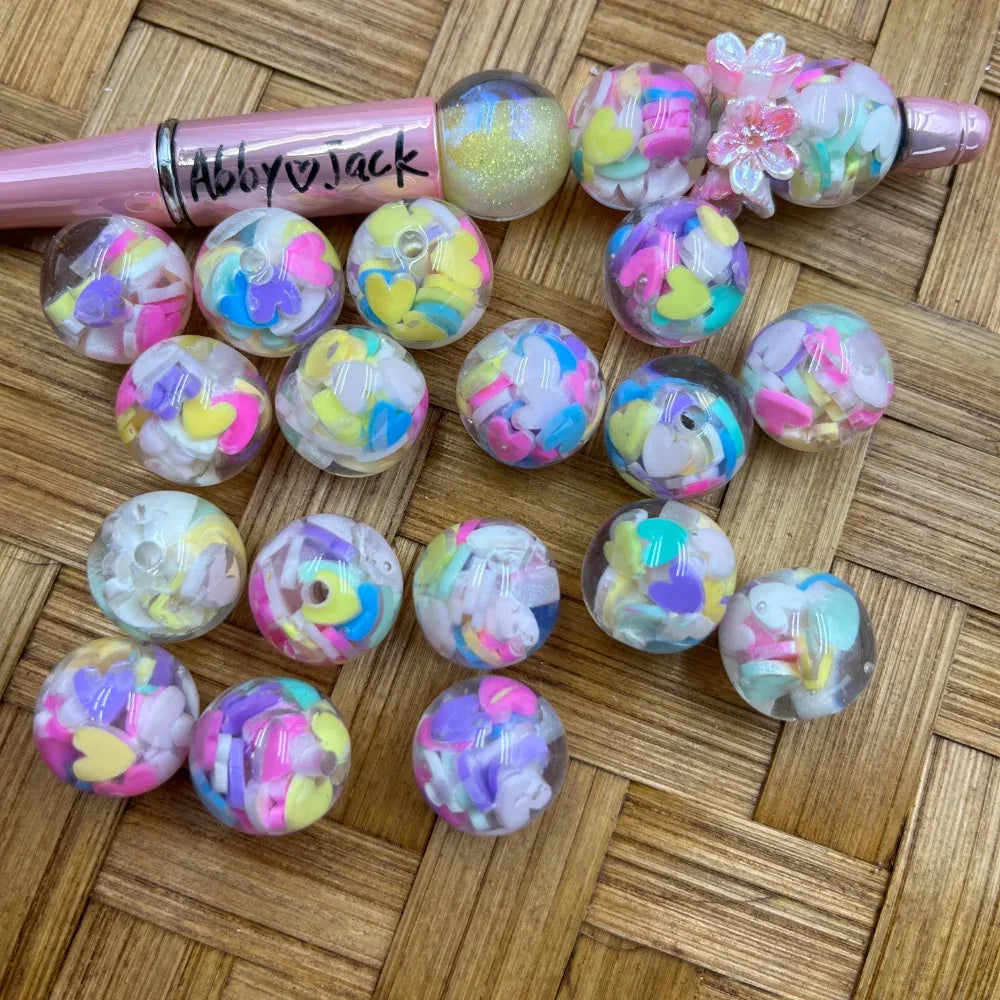 16MM clear round acrylic beads filled with pastel rainbow confetti hearts and flower shapes, displayed next to a pink beadable pen on a wicker surface
