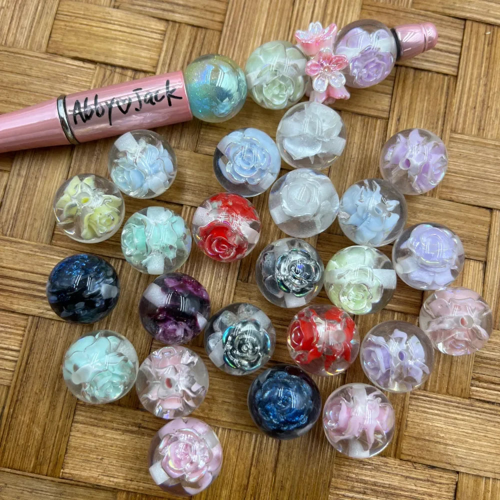 16MM clear acrylic beads with encased 3D rose flowers in assorted colors including red, blue, purple, pink, mint, white, and iridescent, displayed on a woven surface next to a pink beadable pen.