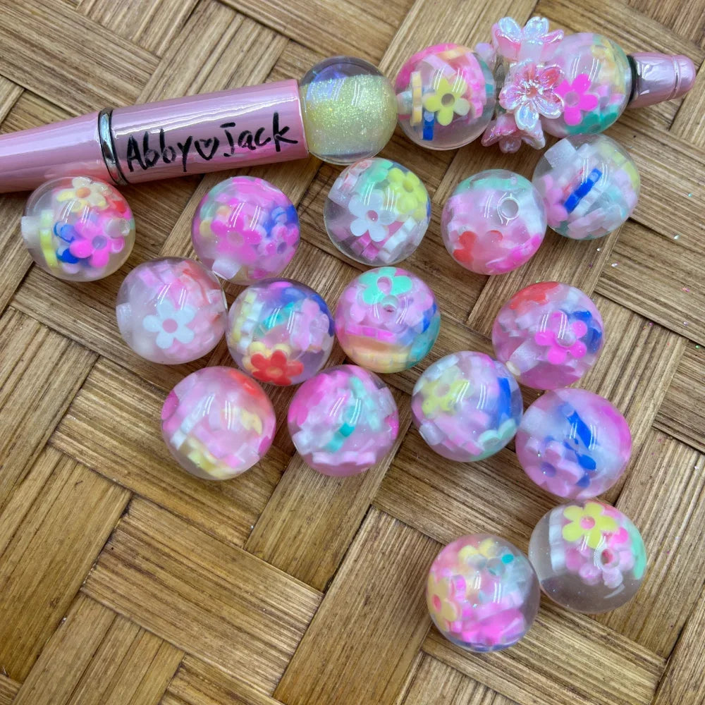 16MM clear acrylic round beads filled with colorful flower and confetti pieces in pink, yellow, blue, and green, displayed next to a pink beadable pen on a wicker surface.