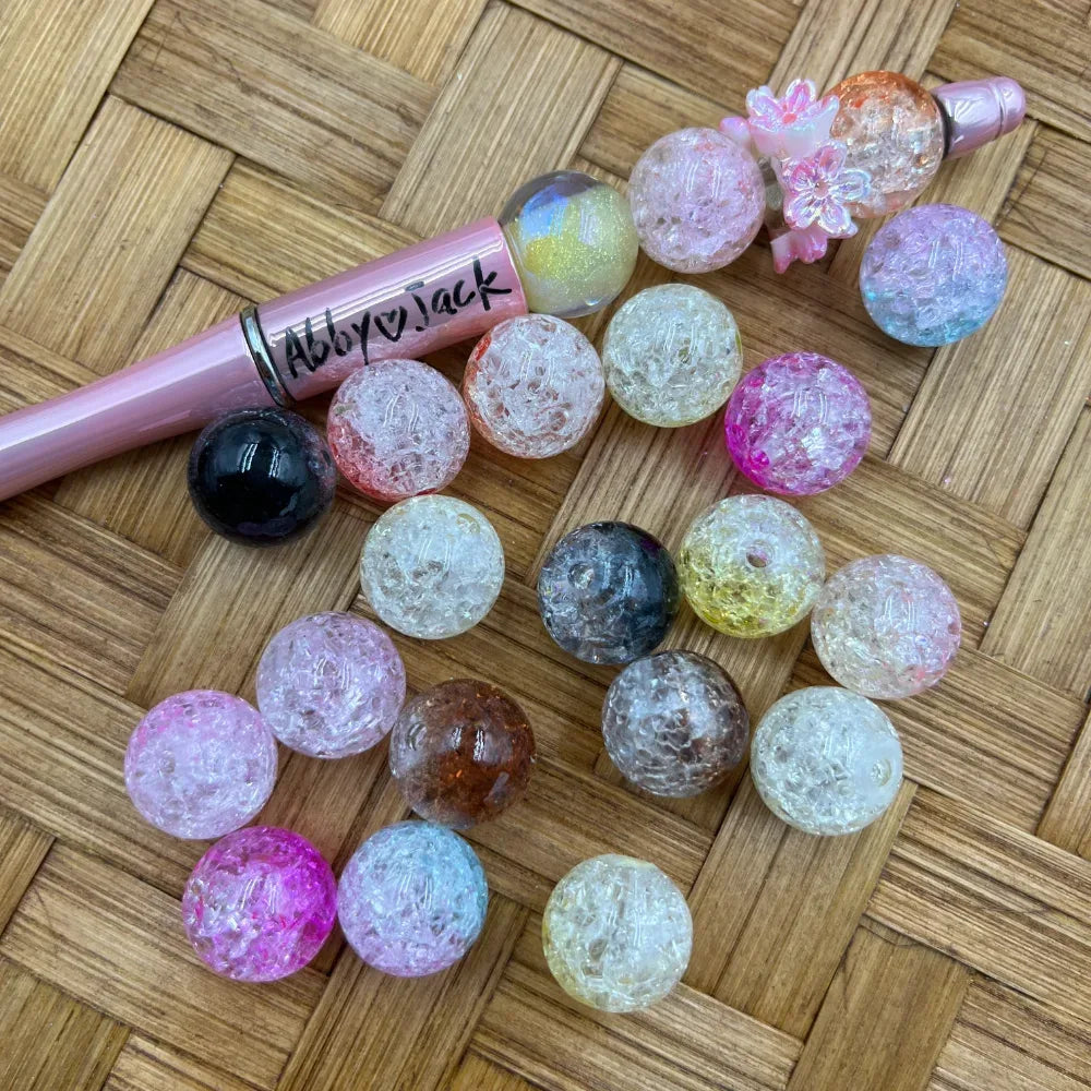 16MM crackle acrylic beads in assorted colors including pink, clear, black, yellow, and blue displayed on a wicker surface next to a pink beadable pen labeled Abby Jack.