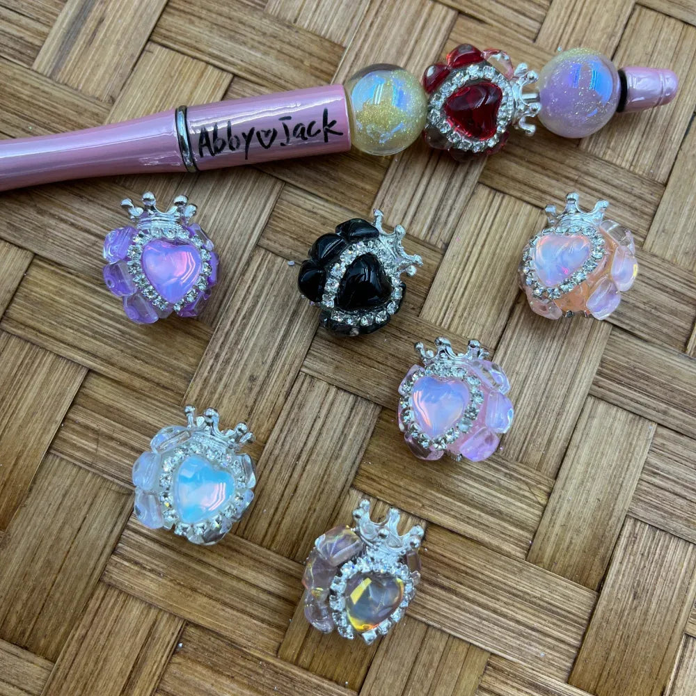 16MM crown heart rhinestone fancy beads in multiple colors including purple, black, pink, white opal, and clear iridescent, displayed on a woven surface next to a decorated beadable pen labeled Abby Jack.