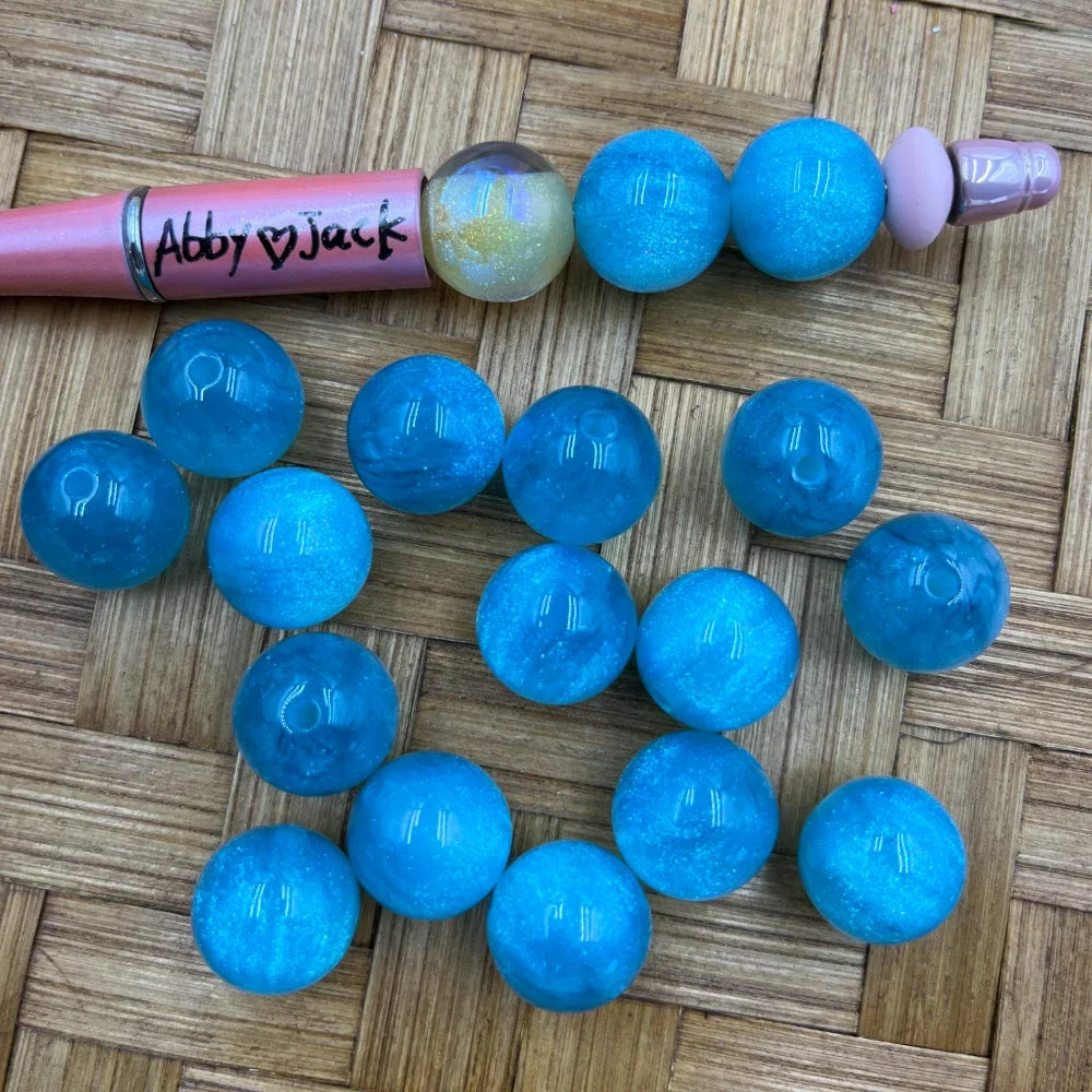16MM cyan blue glitter shimmer acrylic beads displayed on a woven surface next to a pink beadable pen, showing sparkly translucent finish with hole openings visible.
