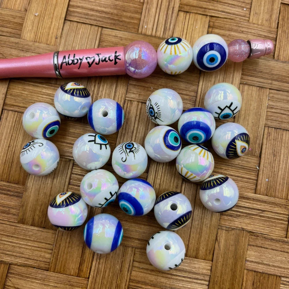 16MM iridescent white acrylic round beads featuring various evil eye designs with blue, teal, and gold accents, scattered on a wicker surface next to a beadable pen labeled Abby and Jack.