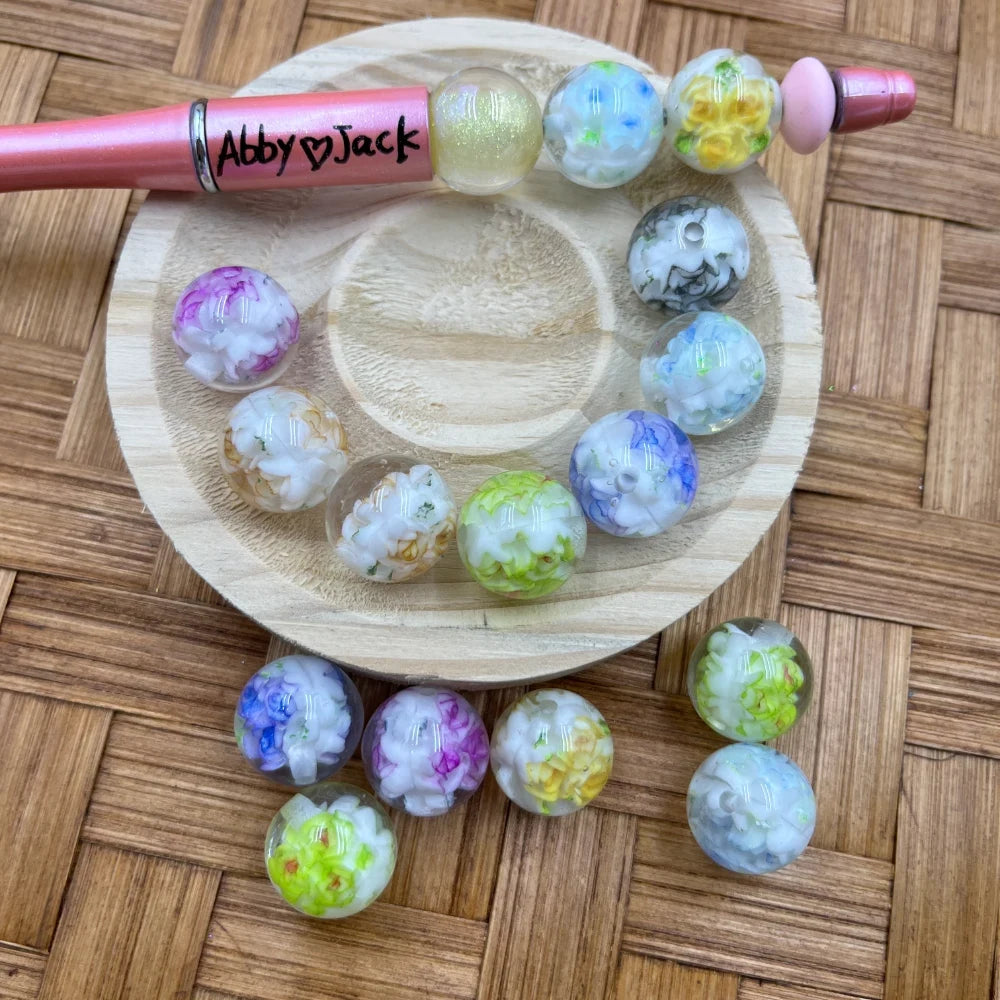 16MM floral flower pattern acrylic beads in assorted pastel colors including purple, pink, yellow, green and blue displayed on a wooden dish with a beadable pen labeled Abby and Jack