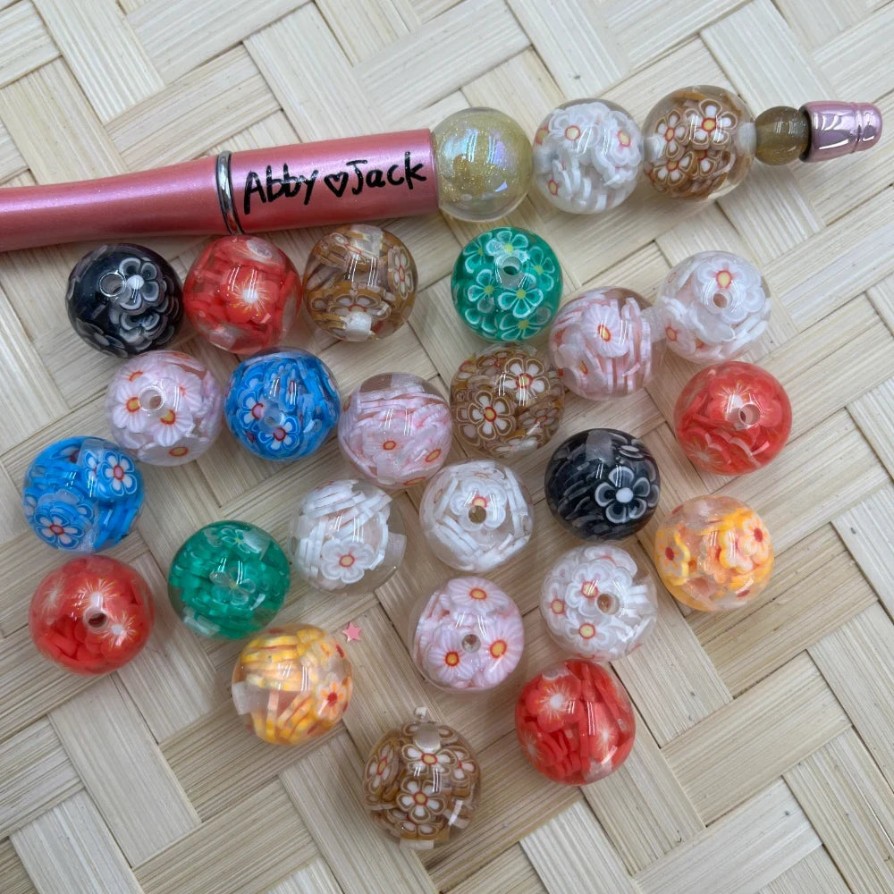16MM floral millefiori acrylic beads in assorted colors including red, black, blue, green, pink, and gold with flower patterns, displayed next to a beadable pen on a wicker surface.
