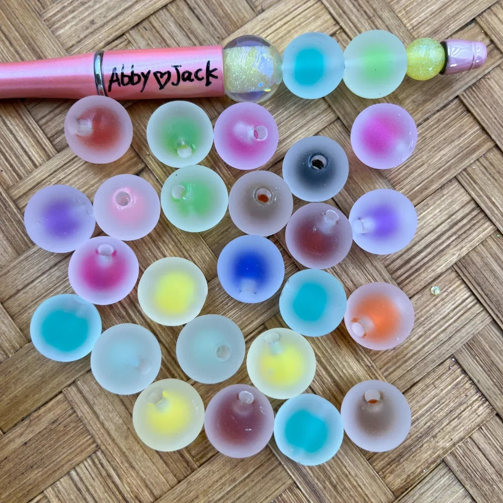 16MM frosted acrylic beads with colorful ombre galaxy center spots in assorted pastel colors including pink, blue, green, yellow, purple, orange and teal, shown with a pink beadable pen on a wicker surface.