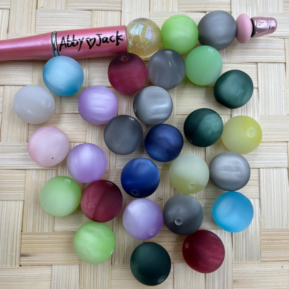 16MM frosted matte cat eye acrylic beads in assorted colors including blue, green, pink, purple, gray, and red arranged on a woven surface next to a beadable pen.
