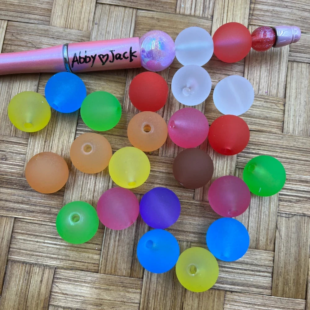 16MM frosted matte multicolor acrylic beads in assorted colors including red, blue, green, yellow, orange, purple, pink, and white, displayed next to a pink beadable pen labeled Abby and Jack on a wicker surface.