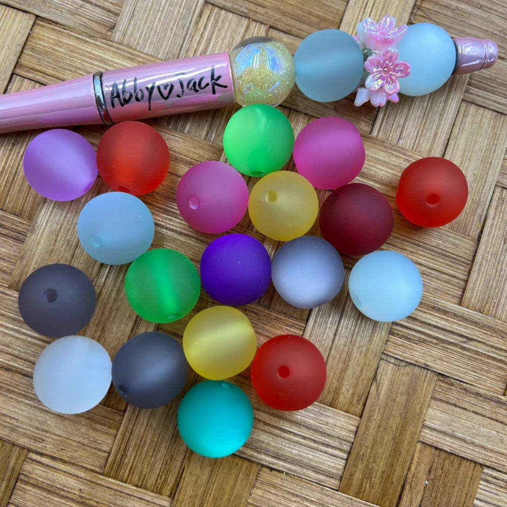16MM frosted matte round acrylic beads in assorted colors including red, green, purple, teal, yellow, pink, gray, white and blue displayed on a wicker surface next to a beadable pen.