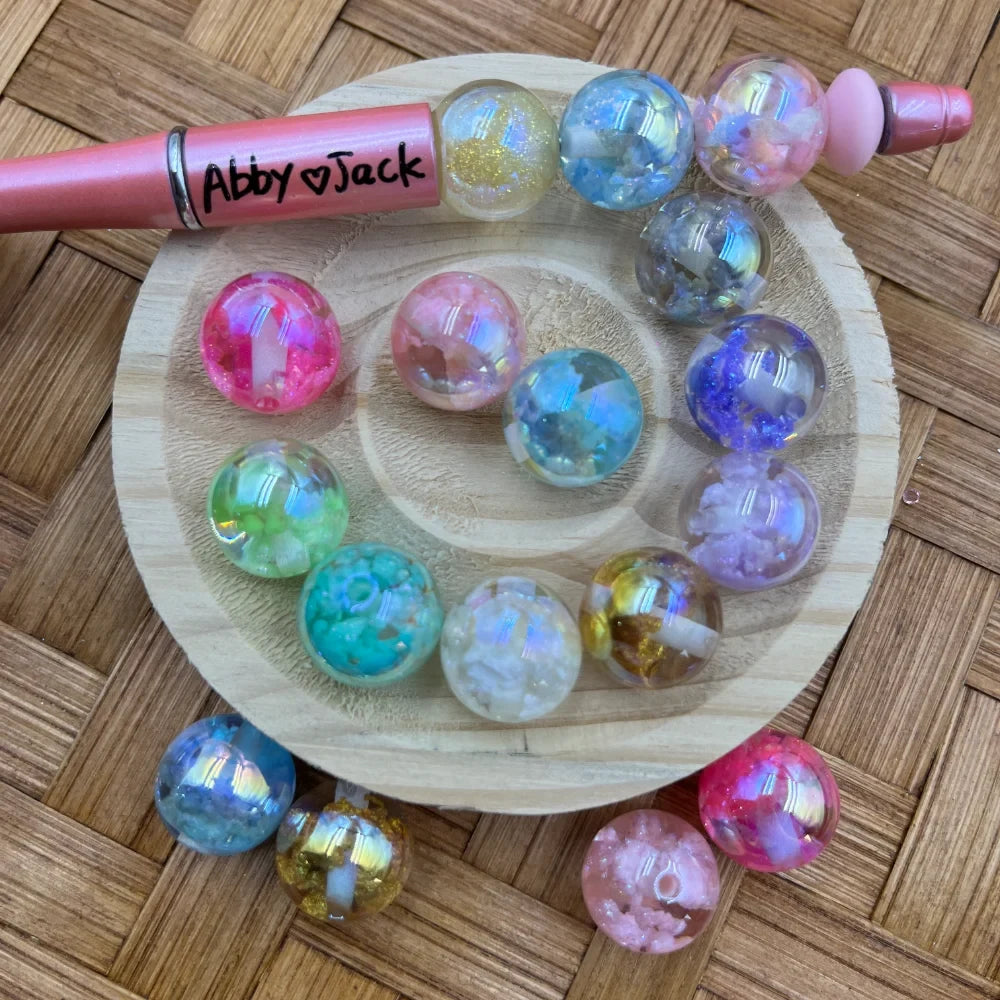 16MM galaxy aurora iridescent acrylic beads in assorted colors including pink, blue, teal, purple, gold, and clear with glitter and crystal fills, displayed on a wooden tray with a beadable pen labeled Abby and Jack.