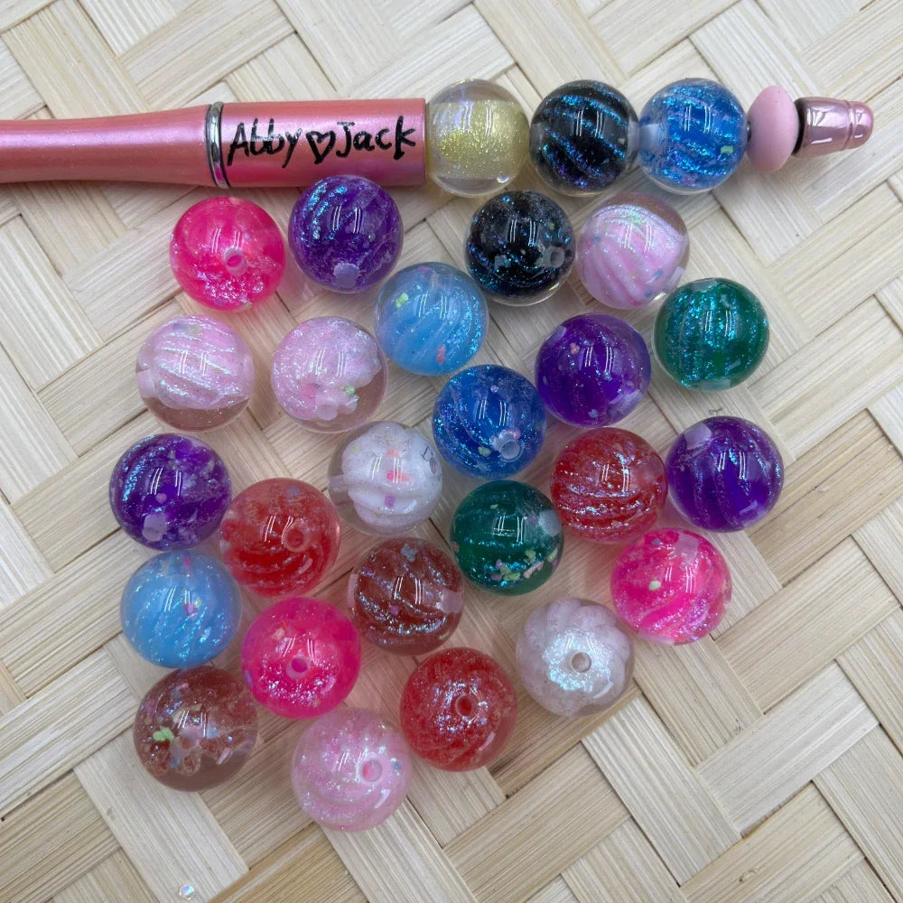 16MM galaxy glitter acrylic beads in mixed colors including pink, purple, blue, teal, black, red and clear with sparkle finish, displayed next to a pink beadable pen on wicker surface.