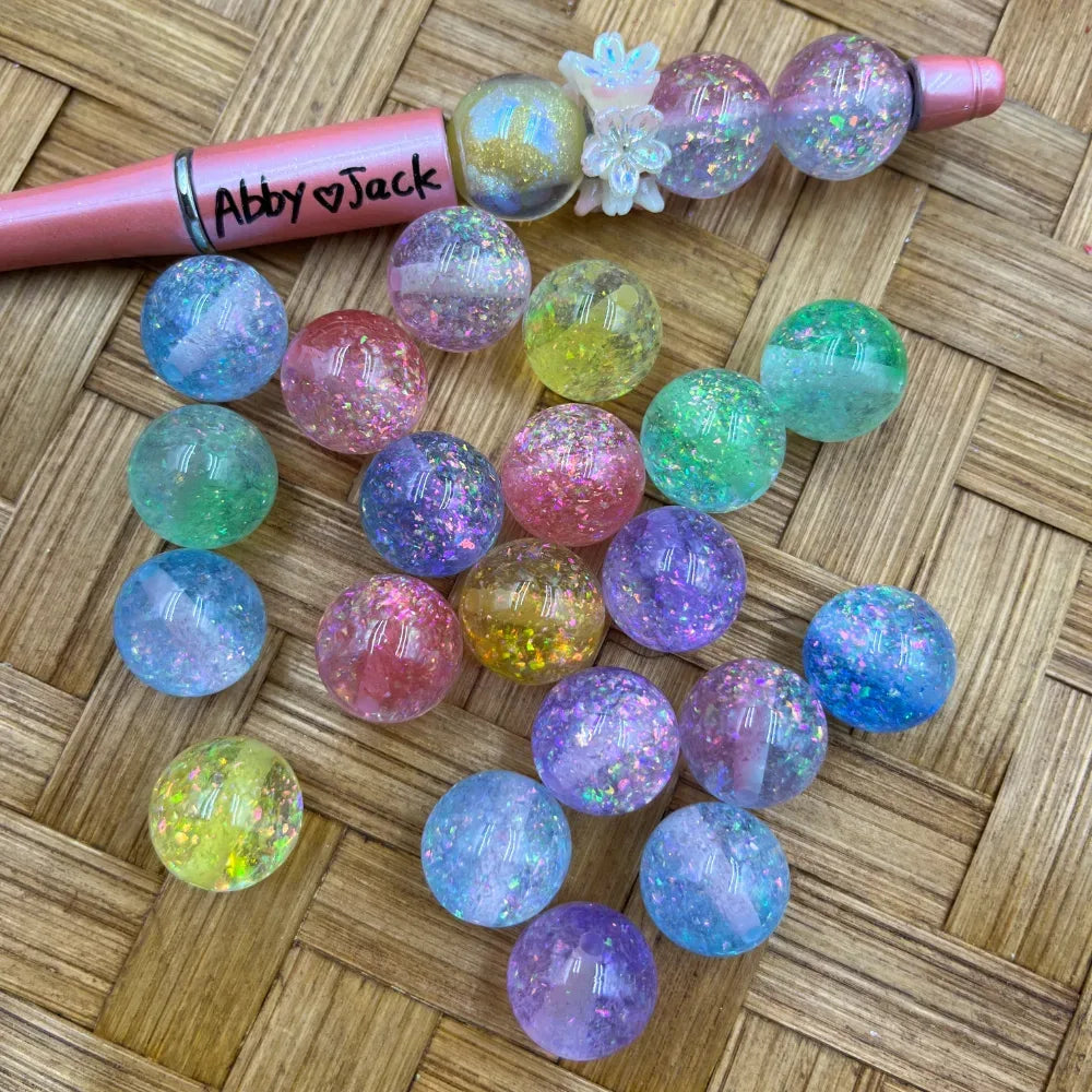 16MM pastel galaxy glitter acrylic beads in assorted colors including pink, blue, green, purple, and yellow with iridescent sparkle flakes, displayed next to a beadable pen on a wicker surface.