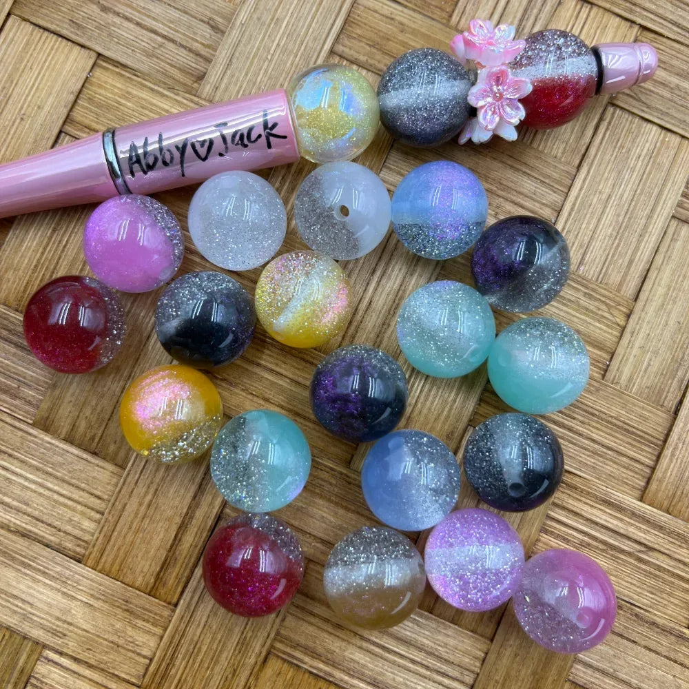 16MM galaxy glitter acrylic beads in assorted two-tone colors including teal, purple, pink, yellow, red, and clear with silver glitter sparkle finish, displayed on woven bamboo surface next to a pink beadable pen.