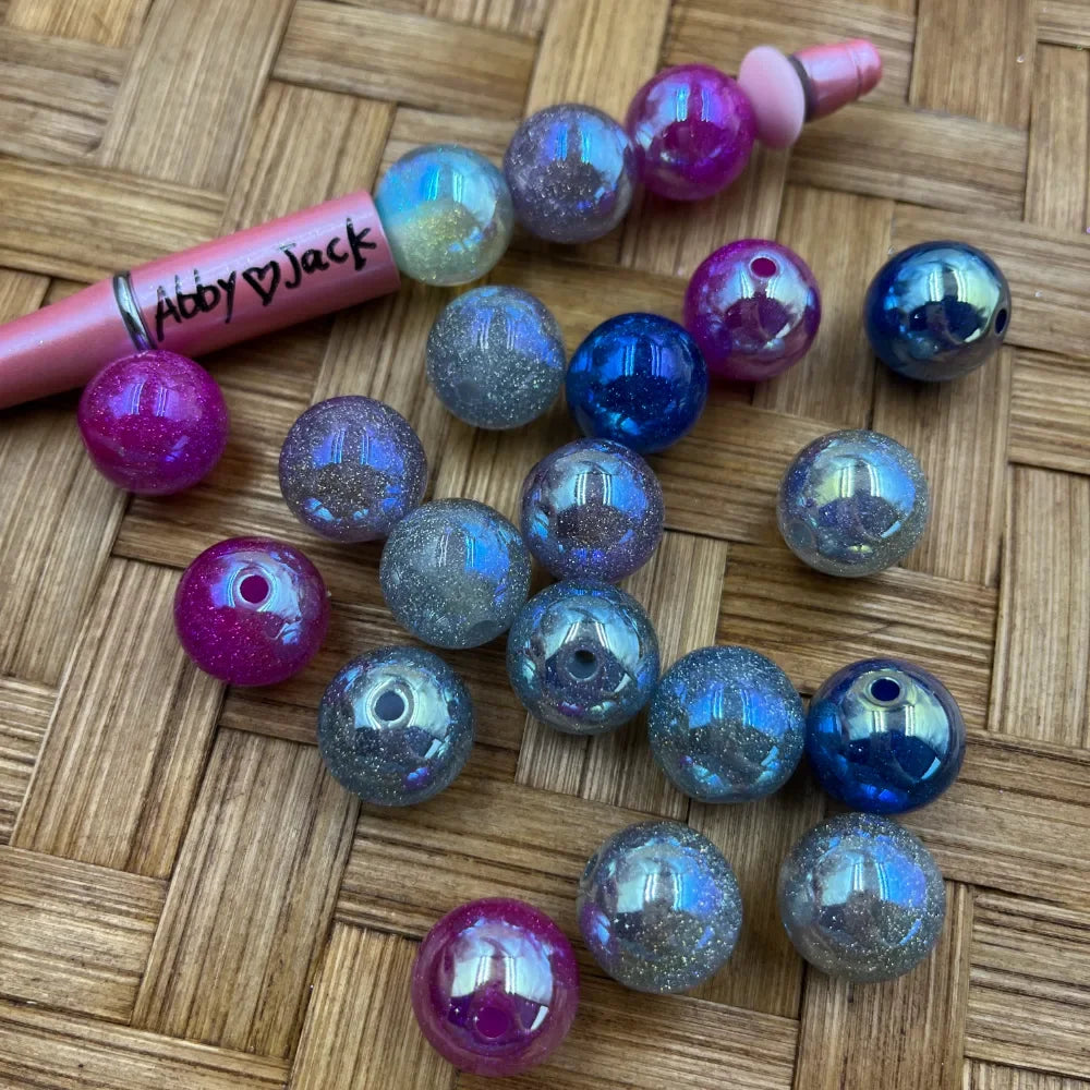 16MM galaxy glitter aurora acrylic beads in iridescent blue, magenta pink, and silver scattered on a woven surface next to a pink beadable pen branded Abby Jack