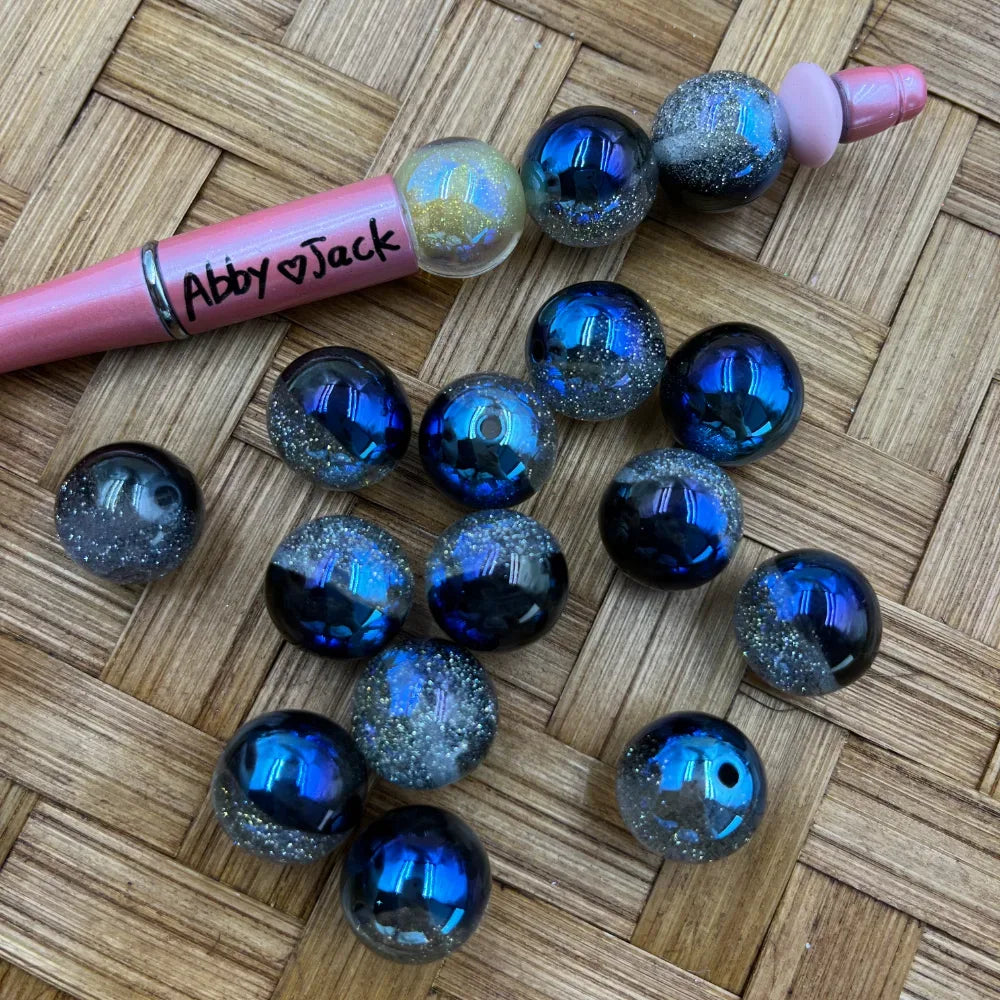 16MM galaxy glitter acrylic beads in deep blue and black with silver glitter sparkle effect, shown with a pink beadable pen on a woven surface