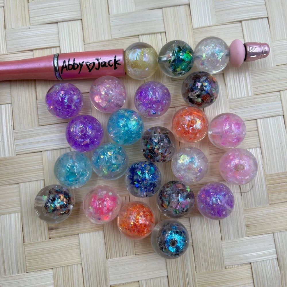 16MM galaxy glitter confetti acrylic beads in assorted colors including purple, teal, pink, orange, and black with holographic sparkle flakes, displayed next to a beadable pen on a wicker surface.