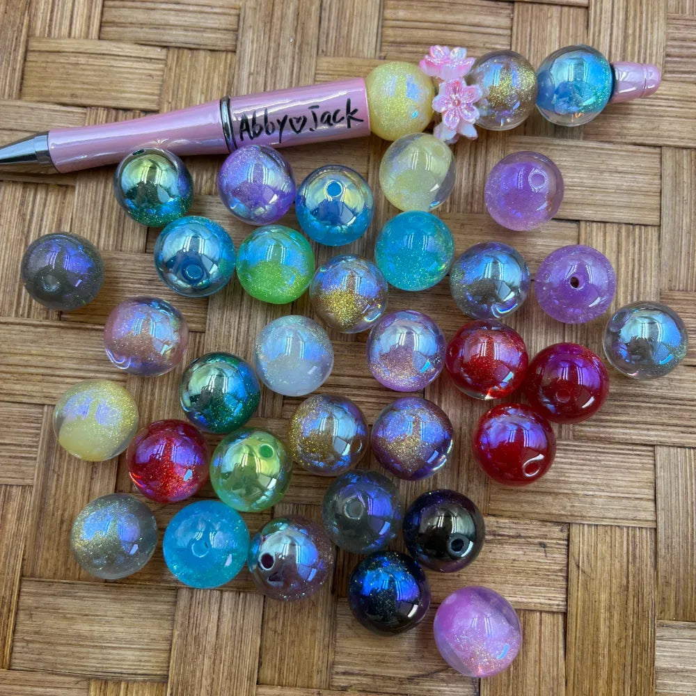 16MM galaxy glitter iridescent acrylic beads in assorted colors including teal, red, purple, green, and pink with aurora shimmer finish, displayed on a woven surface next to a beadable pen.