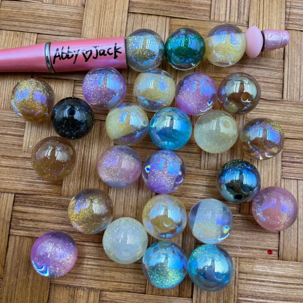 16MM galaxy glitter iridescent acrylic beads in assorted aurora colors including purple, blue, pink, teal, black, and champagne with sparkling glitter finish on woven background next to a pink beadable pen.