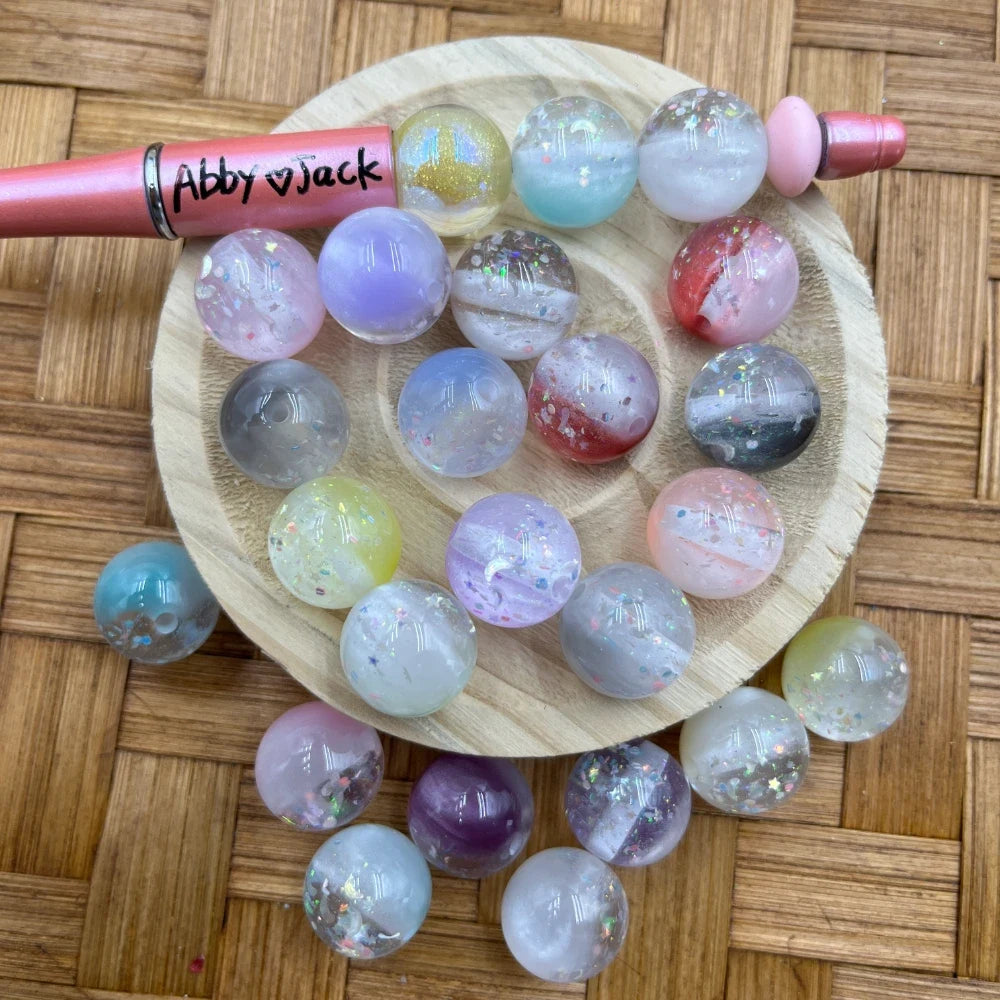 16MM galaxy glitter swirl acrylic beads in assorted pastel and jewel tone colors displayed on a wooden dish next to a beadable pen, showing iridescent glitter and marbled swirl patterns