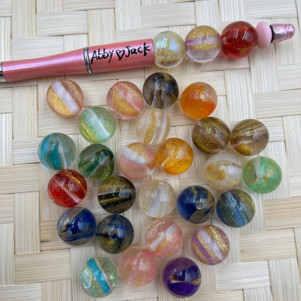 16MM multicolor galaxy glitter swirl acrylic beads in assorted colors including teal, red, blue, pink, green and gold shimmer, displayed next to a pink beadable pen on a woven surface.