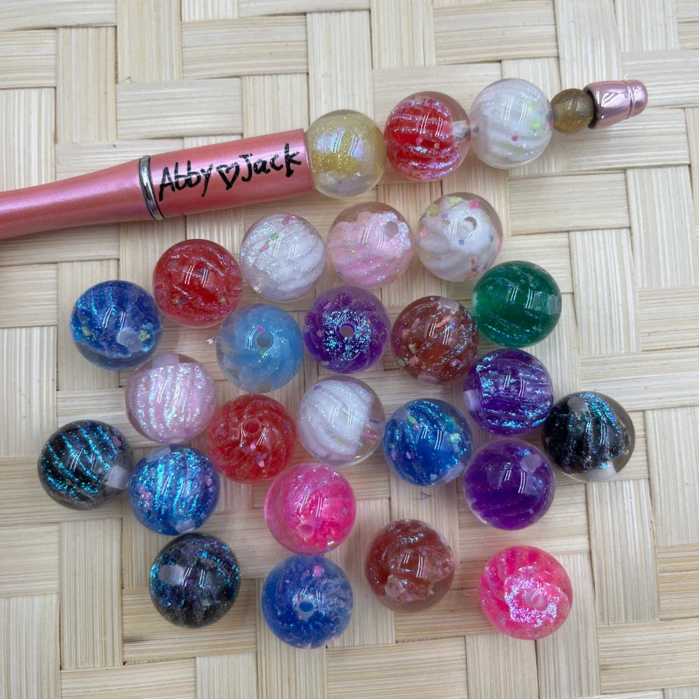 16MM galaxy glitter acrylic beads in assorted colors including blue, red, pink, purple, white, and black with sparkle swirl effects, displayed beside a beadable pen on a wicker surface.