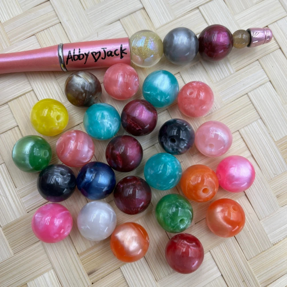 16MM galaxy marble acrylic beads in assorted pearlescent colors including teal, pink, burgundy, orange, navy, and more, shown loose on a woven surface next to a beaded pen labeled Abby Jack.