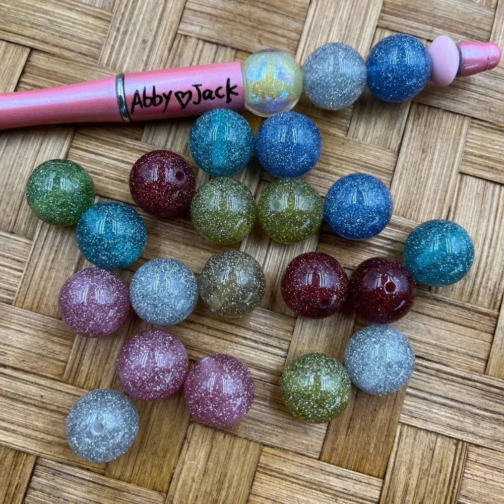 16MM glitter acrylic beads in assorted colors including blue, teal, red, green, pink, clear, and gold sparkle, displayed next to a pink beadable pen on a wicker surface.