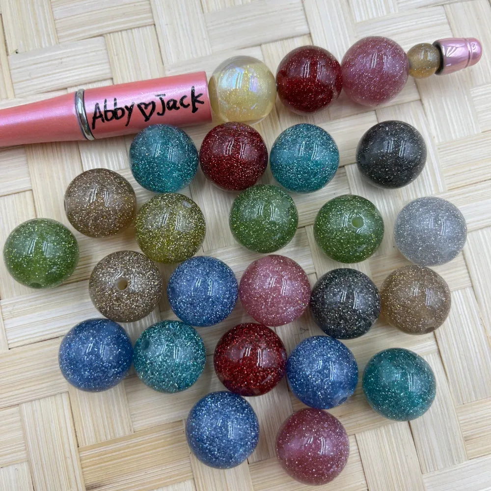 16MM glitter acrylic beads in assorted sparkle colors including blue, pink, green, red, teal, gold, and gray displayed on a wicker surface next to a beadable pen.