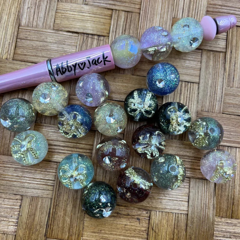 16MM round acrylic beads with gold bow charms and glitter finish in multiple colors including blue, black, green, purple, and clear, displayed next to a pink beadable pen on a wicker surface.