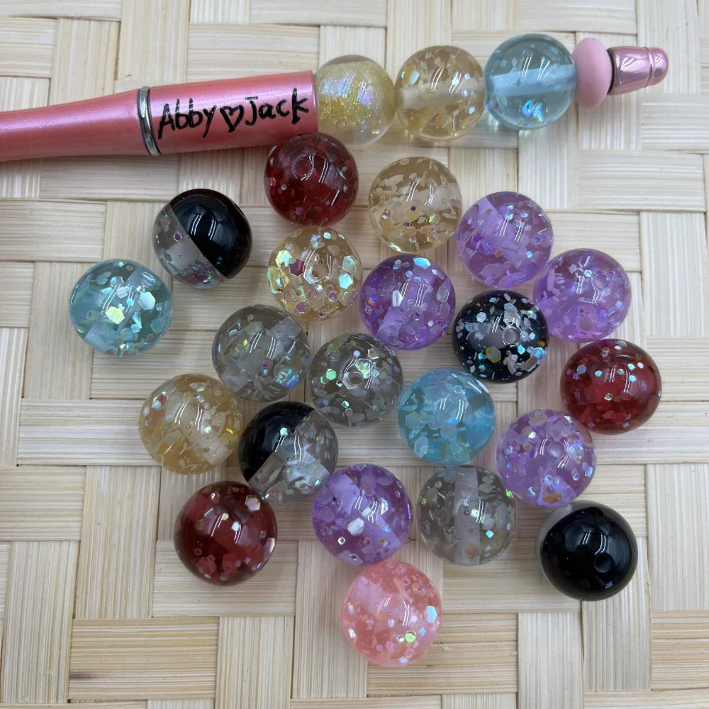 16MM glitter confetti acrylic beads in assorted colors including purple, black, red, blue, pink, clear, and gold displayed next to a pink beadable pen on a woven surface.