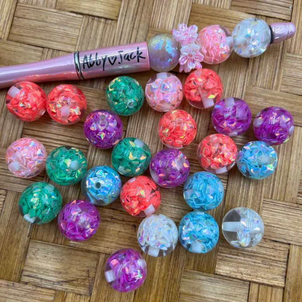 16MM iridescent glitter confetti acrylic beads in assorted colors including coral, teal, purple, pink, and white, displayed next to a pink beadable pen on a woven surface.