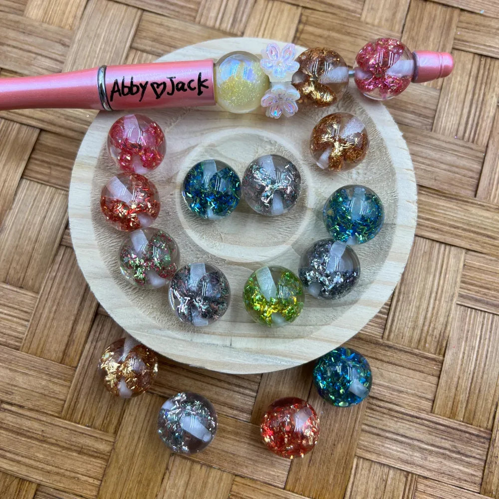 16MM round acrylic beads with glitter foil and cross inclusions in multiple colors including pink, teal, gold, green, and silver, displayed on a wooden tray with a beadable pen.