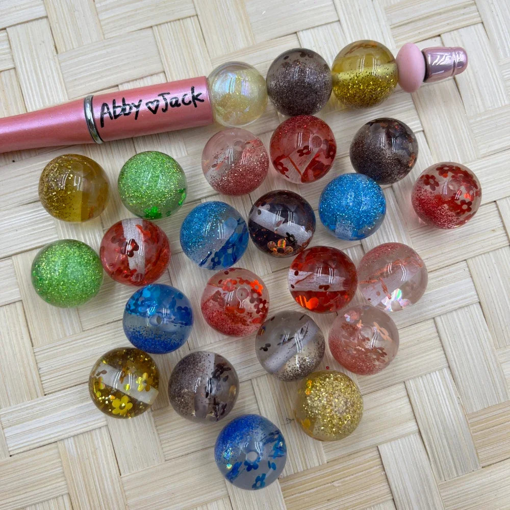 16MM glitter galaxy acrylic beads in assorted colors including red, blue, green, gold, and clear with floral confetti and sparkle inclusions, displayed on woven surface next to a pink beadable pen.