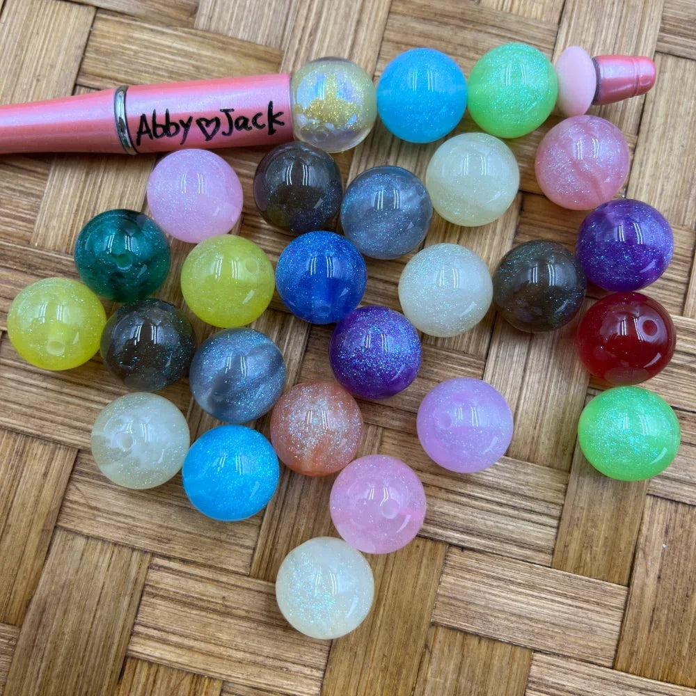 Assorted 16MM glitter galaxy acrylic beads in multiple colors including purple, blue, green, pink, red, yellow, and white, displayed next to a pink beadable pen on a wicker surface.