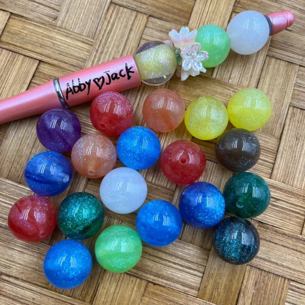 16MM assorted glitter galaxy acrylic beads in multiple colors including blue, green, red, purple, teal, yellow, and white, displayed next to a beadable pen on a wicker surface.