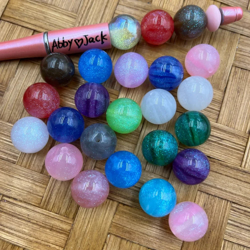 16MM glitter galaxy acrylic beads in assorted colors including blue, pink, green, purple, teal, red, white, and gray displayed on a woven surface next to a pink beadable pen labeled Abby and Jack.